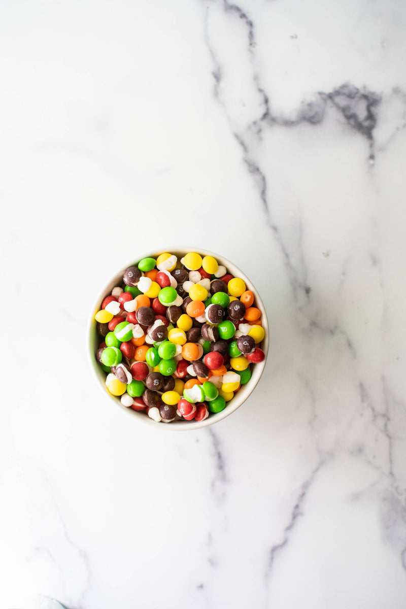 A white bowl filled with colorful, crunchy Freeze Dried Skittles from Sow Good Inc. sits on a marbled surface.