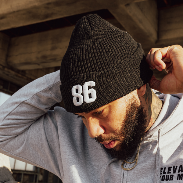 The man adjusts his black beanie with "86," seamlessly paired with a gray hoodie and gold chain—an understated nod to Everyday Delta's Mystery Swag, offering merchandise that's both surprising and stylish.